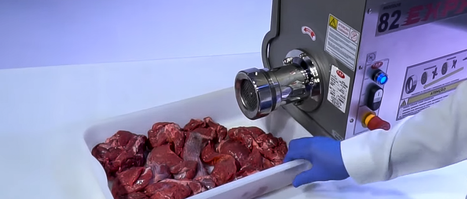 Operador com luva azul posiciona bandeja com pedaços de carne crua em frente a um moedor industrial CAF modelo 82 EXPRESS